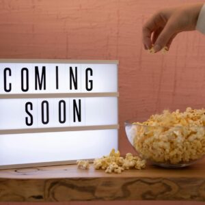 A hand reaching for popcorn next to an illuminated 'Coming Soon' sign, creating a cinematic feel.